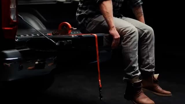 mies istuu pick-up auton lavalla ja vieressä lavalta roikkuu kuormahihna